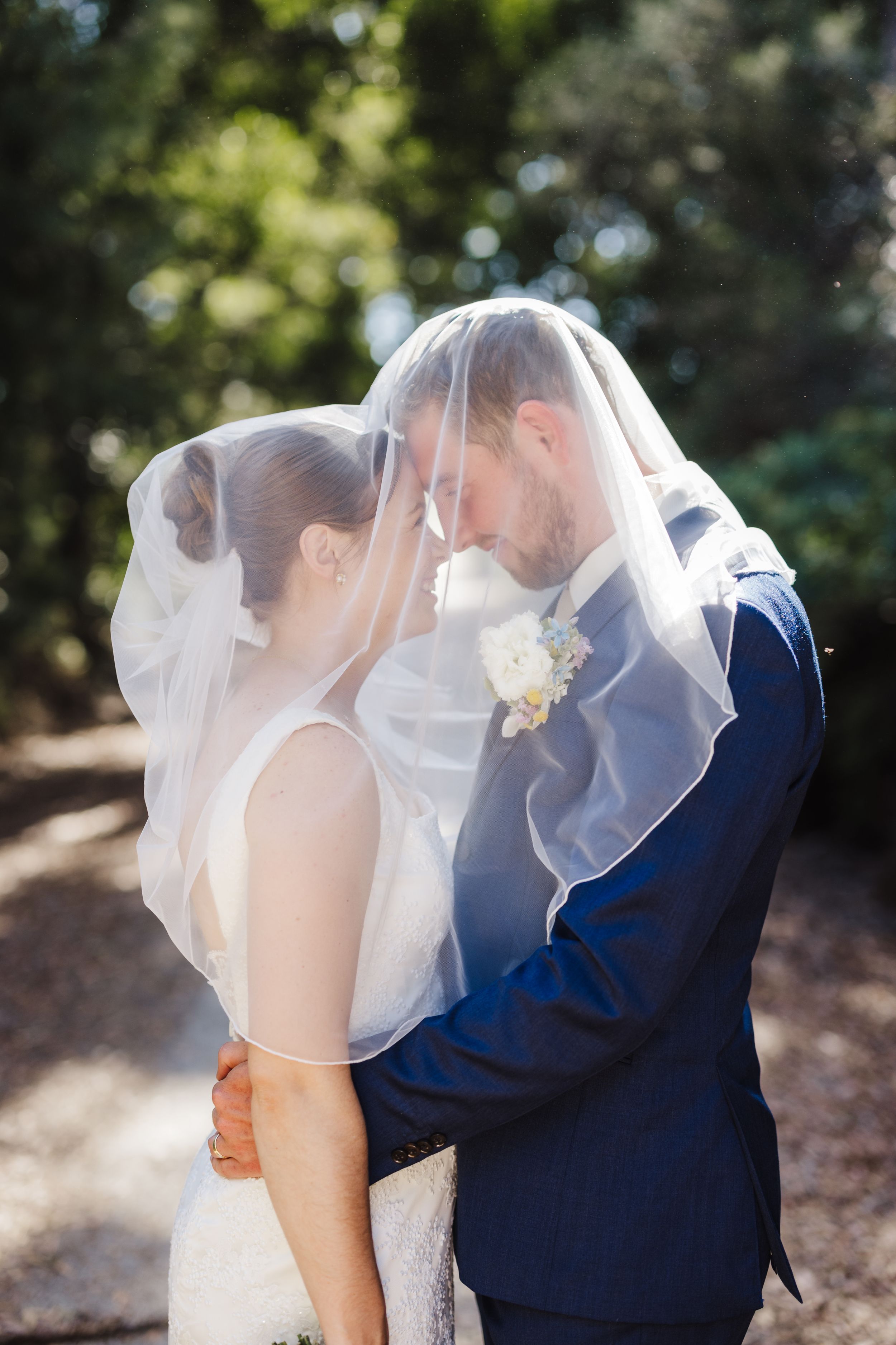 Romantic wedding portrait with couple sharing intimate moment under flowing veil in sunlit garden.