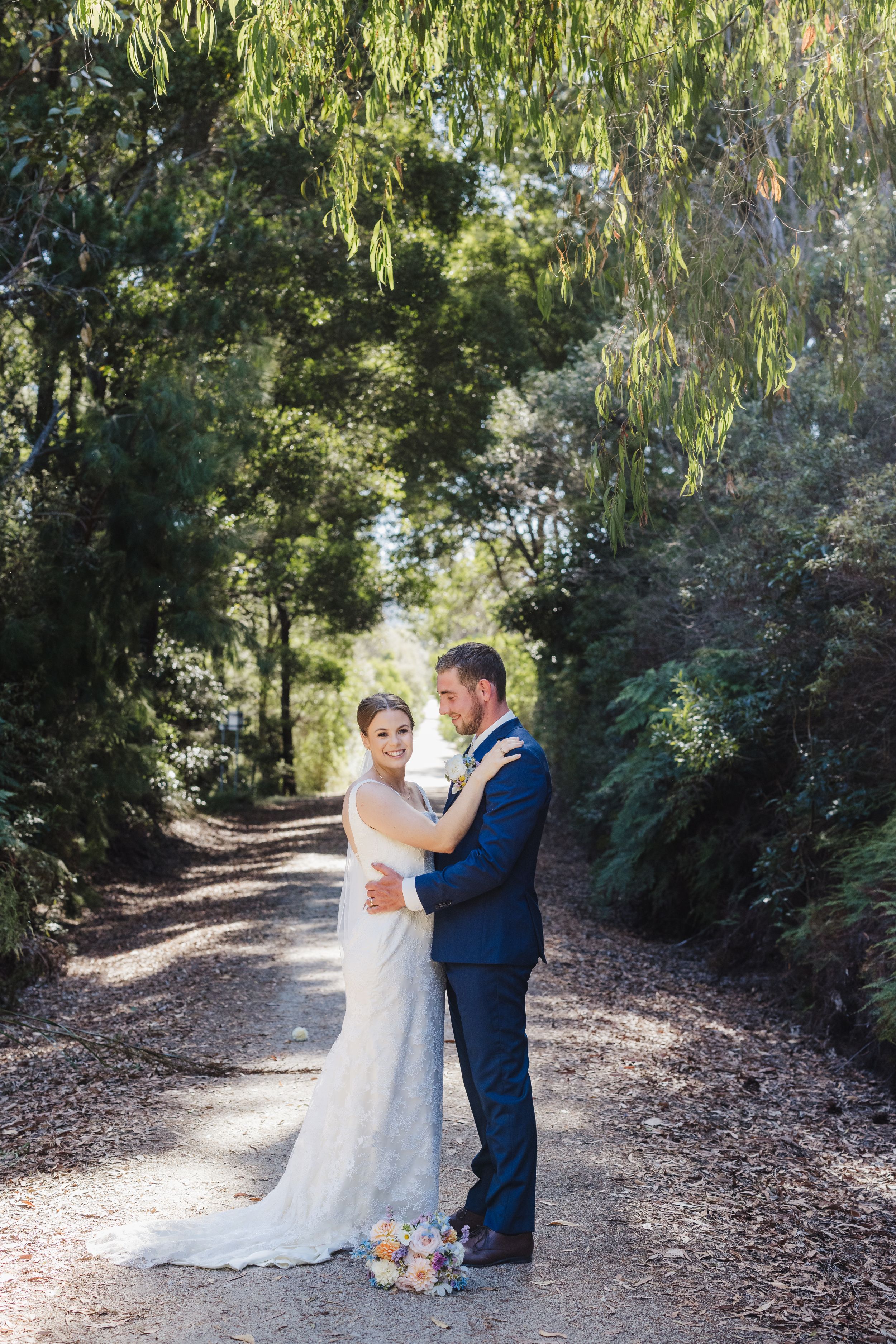 Wedding couple embracing on tree-lined garden path in natural sunlight.