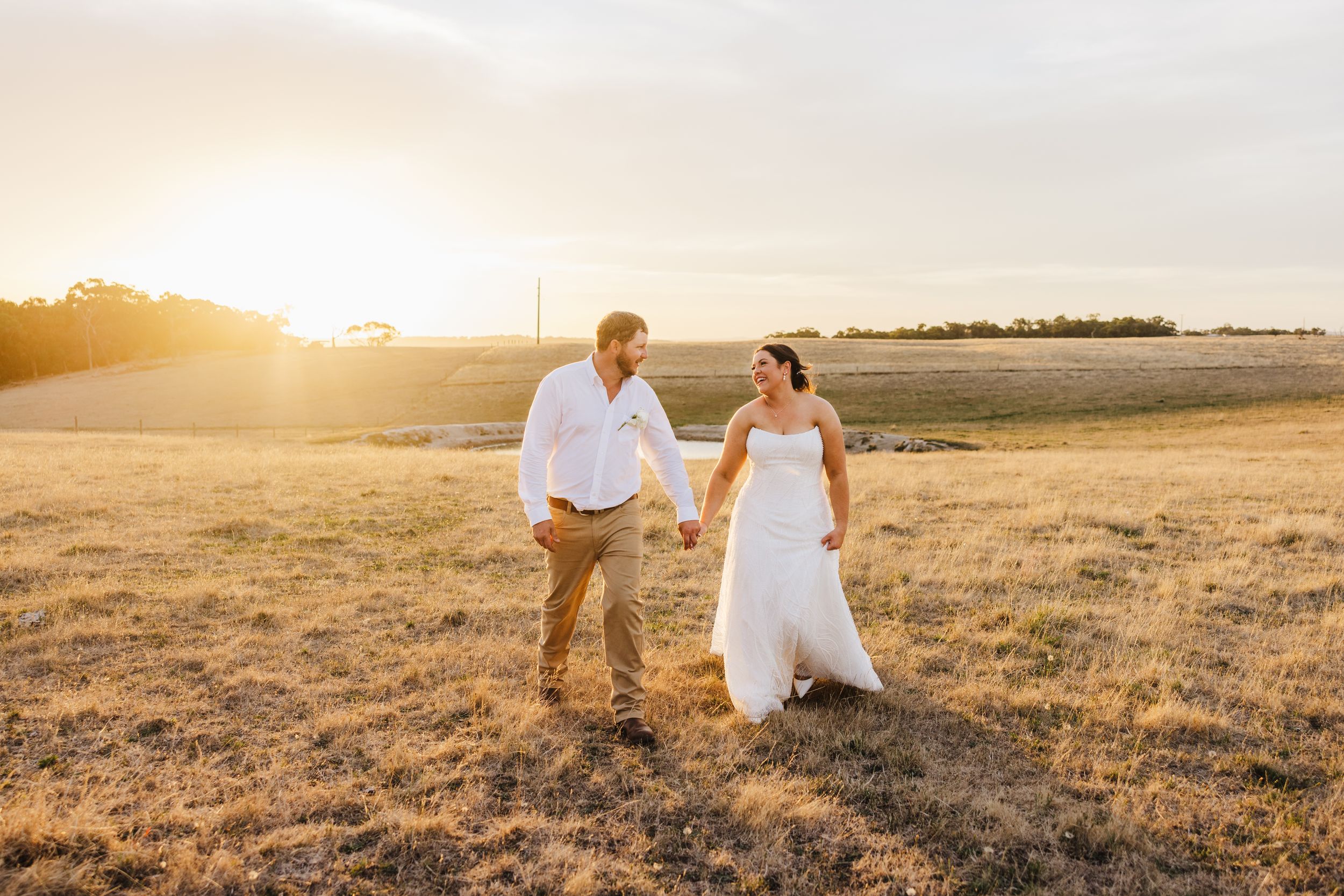 A couple walking together in a golden sunlit field at sunset wearing wedding attire.