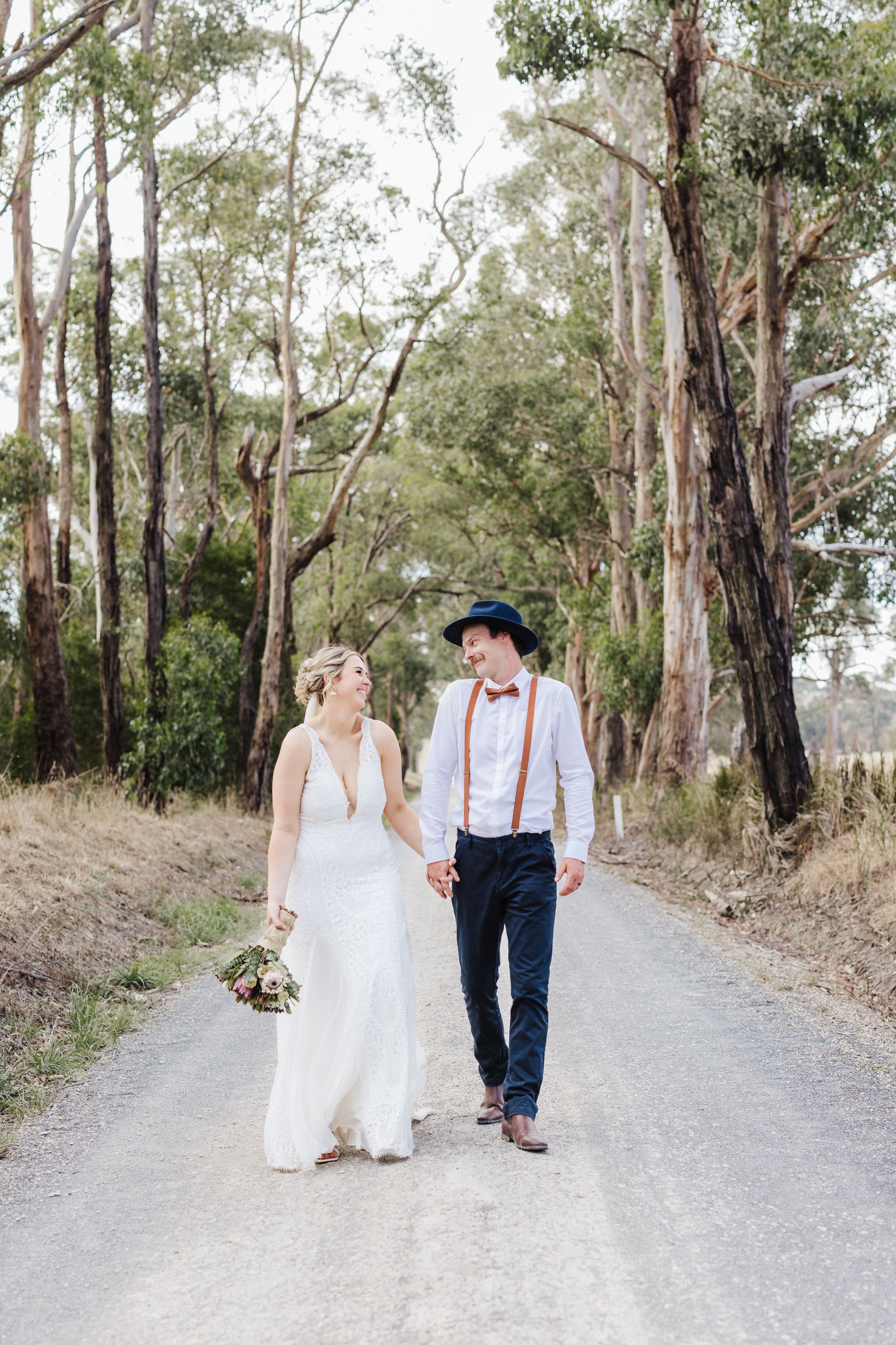 A couple walking together on a rural path lined with tall eucalyptus trees, the bride in white and groom in suspenders.