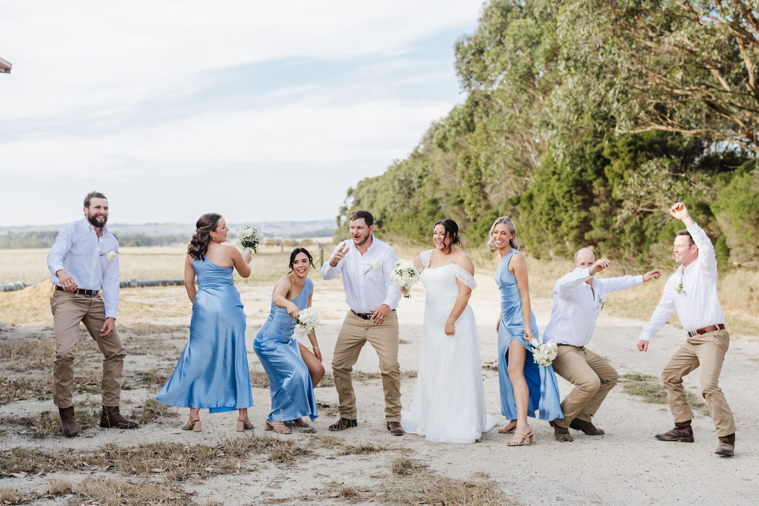 A wedding party strikes playful poses together on a scenic coastal bluff during a casual daytime celebration.