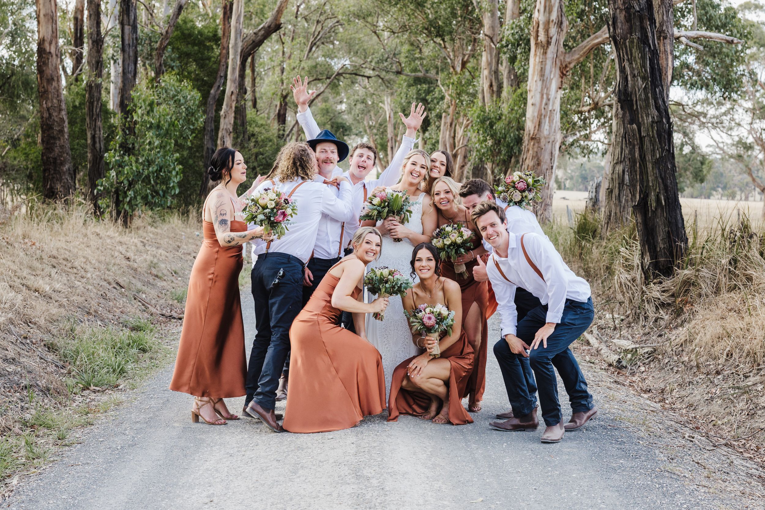 A fun group of wedding party members pose playfully on a dirt road surrounded by tall eucalyptus trees.