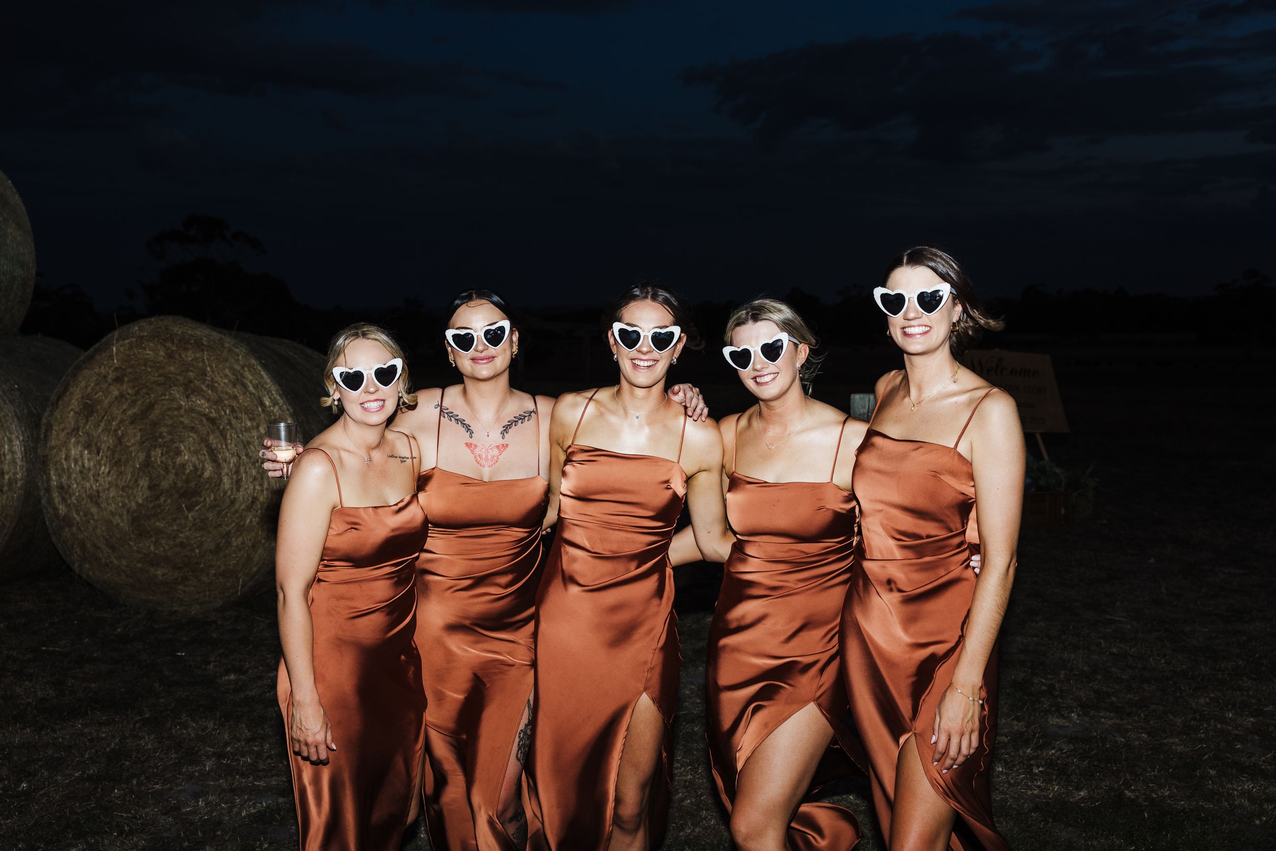 Five bridesmaids in matching rust-colored satin dresses and sunglasses pose together at dusk with hay bales in background.