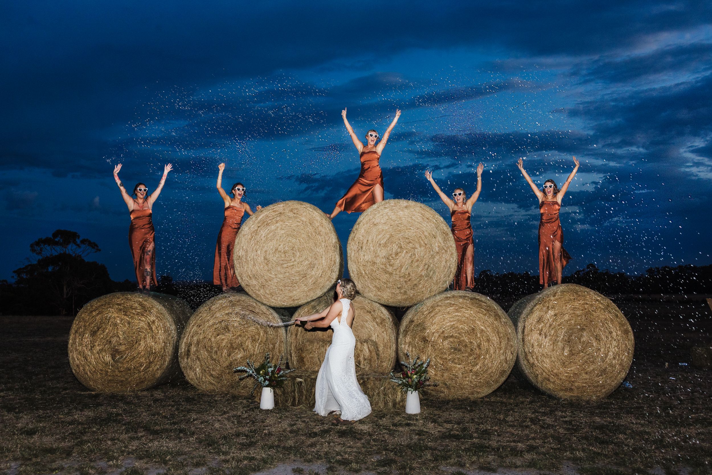 Wedding party poses on hay bales against dramatic night sky with lanterns and rustic farmland backdrop.
