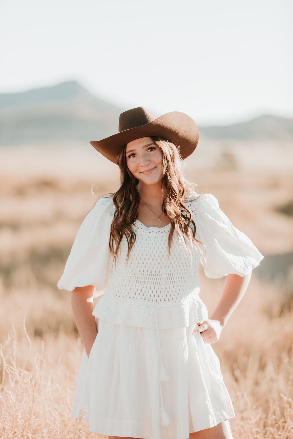 High school senior girl in white lace dress and brown hat poses in sunlit field of golden grass at Little Sahara Sand dunes.