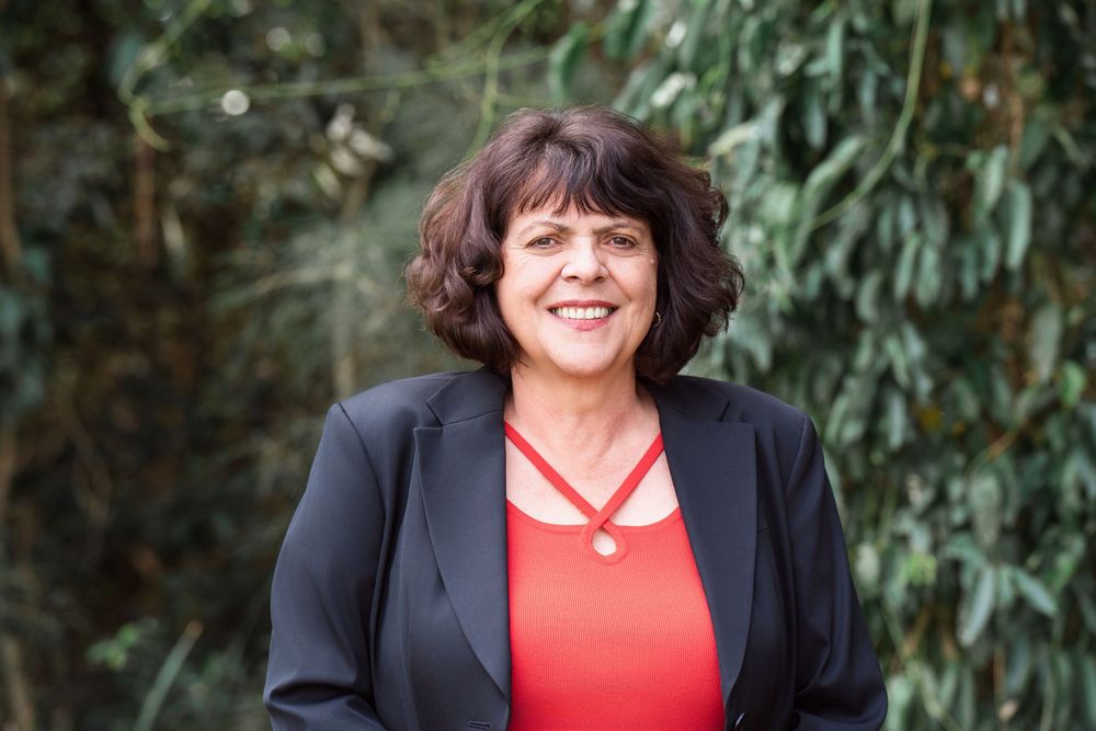 Female outdoors headshot wearing business suit jacket and red top, taken against a soft creamy greenery backdrop of trees.