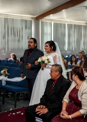 Wedding ceremony in a church hall with blue pews and vertical blinds.