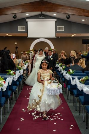 Wedding ceremony procession on burgundy carpet aisle with white decorative arch and flower petals.