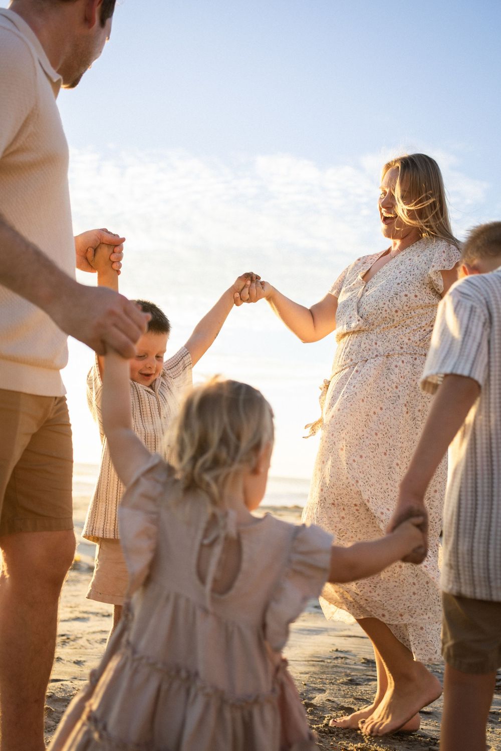 Family holding hands in a circle on a sunny beach at golden hour, wearing neutral tones, children laughing joyfully.