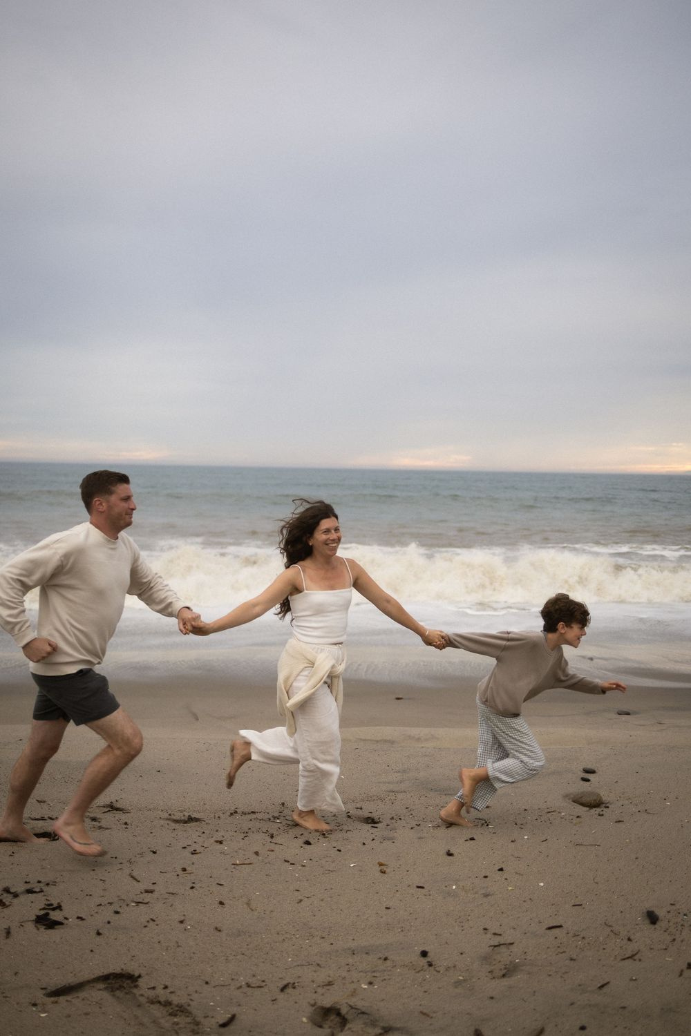 Family of three holding hands and running joyfully on a sandy beach with ocean waves in the background.