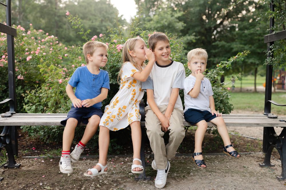 Father sitting on park bench surrounded by three young children in a rose garden setting.