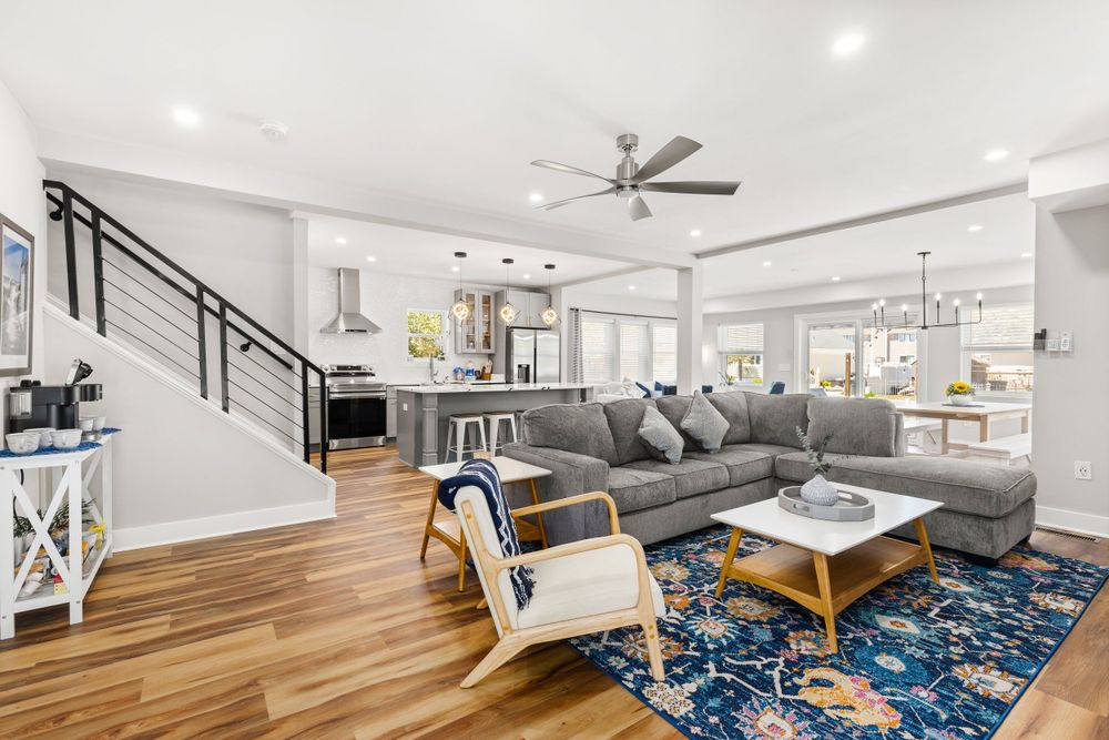 Open-concept living room with gray sectional, hardwood floors, blue rug, and modern staircase.