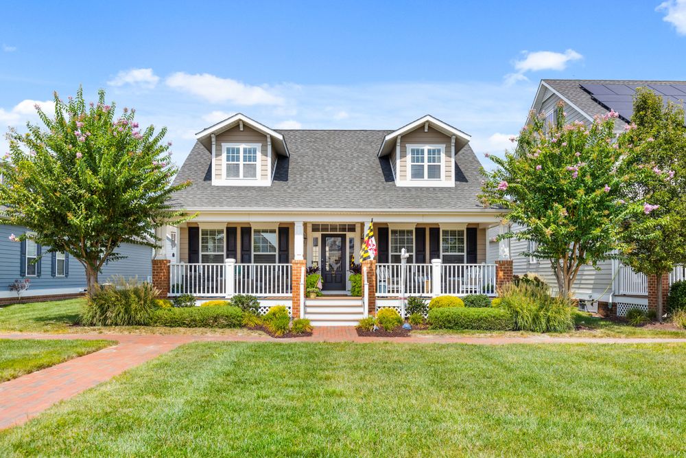 Charming Cape Cod home with covered front porch, dormer windows, and lush green lawn surrounded by mature trees.