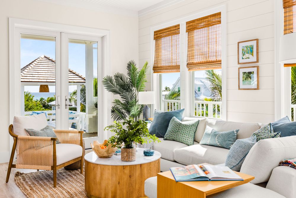 Airy coastal living room with white shiplap walls, bamboo shades, blue pillows, and tropical plants.