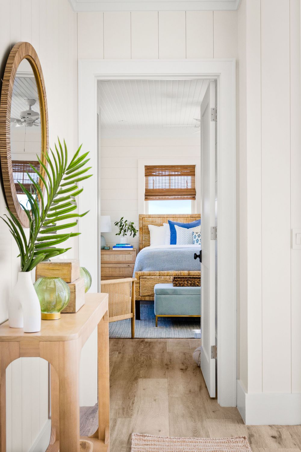 Bright coastal hallway leading to a beachy bedroom with rattan furniture, woven shades, and blue accents.
