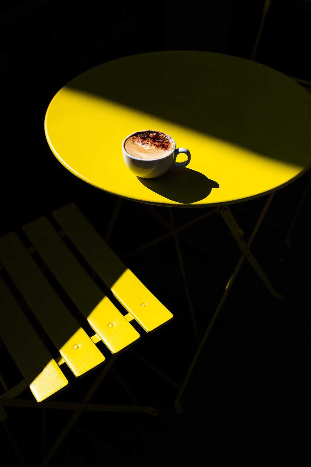 Cup of cappuccino on a bright yellow café table, captured in dramatic sunlight with bold shadows and minimalist composition.