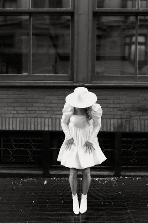 Black and white portrait of person in white dress and hat facing brick building.