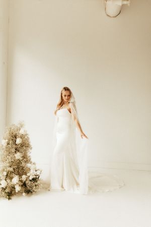 Ethereal bridal portrait with elegant white gown and floral arrangement in minimalist studio.