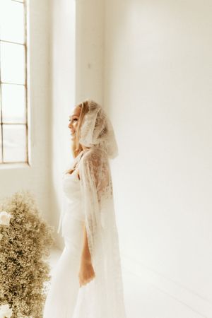 Romantic bridal portrait with lace veil against white wall and dried flower arrangement.