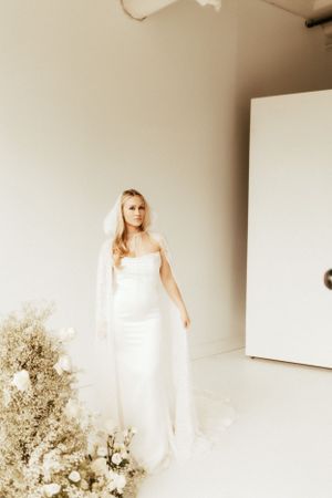 Full-length view of bride in fitted white gown with flowing veil beside floral installation.