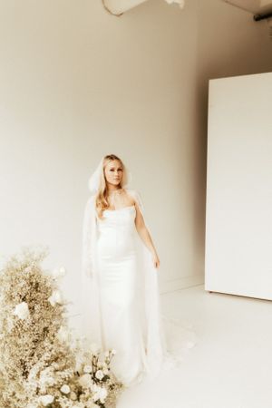 Bride in minimalist white gown poses elegantly beside dried floral arrangement.