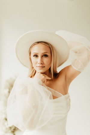 Ethereal bridal portrait featuring a white wide-brim hat and flowing dress against a light background.