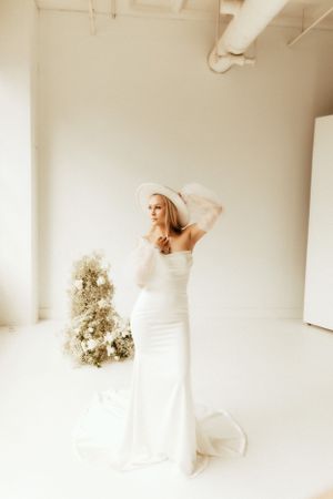 Bride in fitted white gown poses elegantly in minimalist white studio with dried floral arrangement.