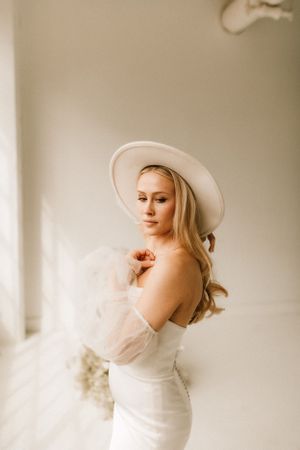 Artistic bridal portrait with white hat and off-shoulder gown against neutral backdrop.
