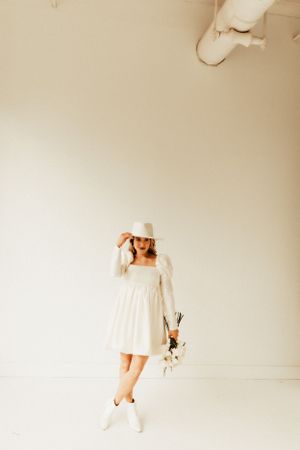 A woman in a white dress and hat poses gracefully in a minimalist white studio setting.