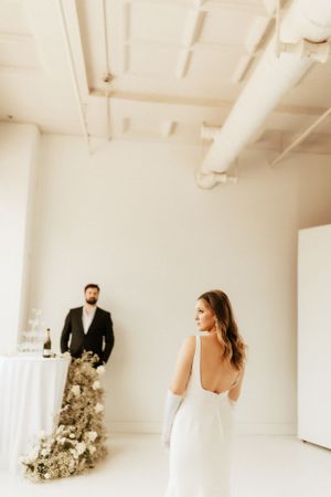 Wedding scene in white room with bride in backless dress and groom standing by floral arrangement.