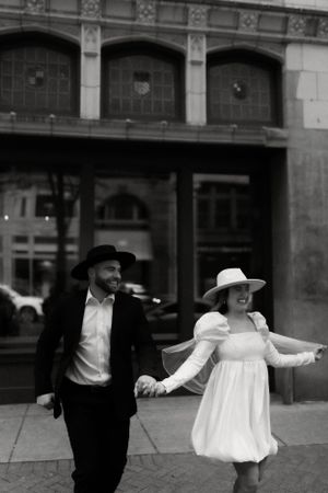 Black and white photo of couple in contrasting outfits walking playfully on city street.