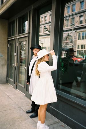 Couple in vintage attire poses on urban street near storefront windows.