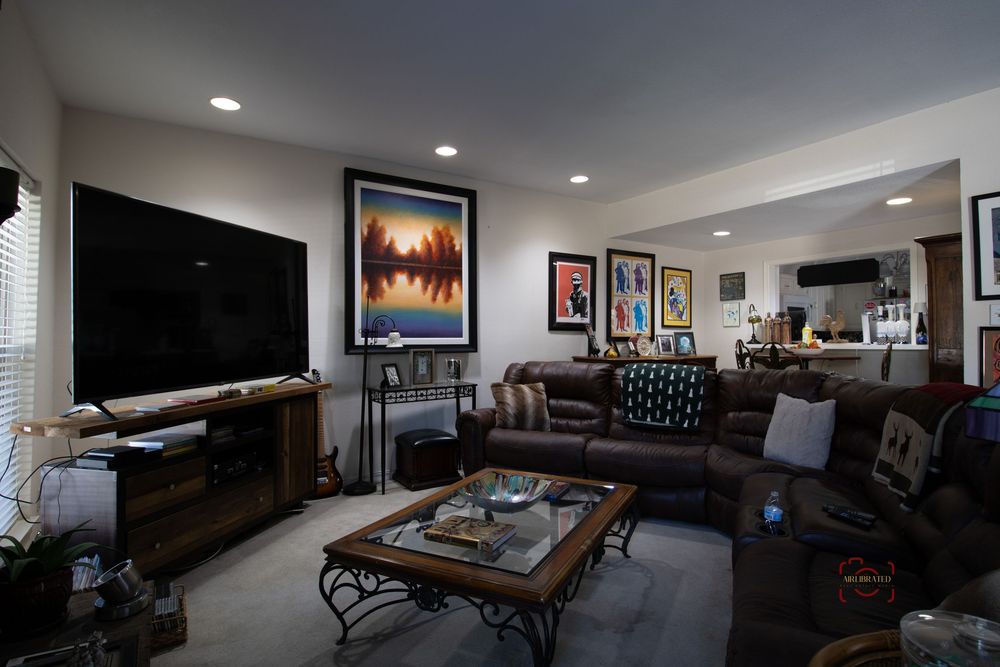 Cozy living room with large flat-screen TV, dark leather sectional sofa, coffee table, and colorful artwork on the walls.