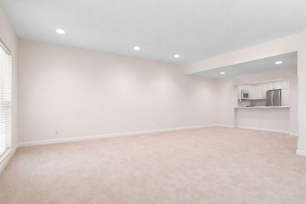 Bright, open living room with plush carpet, recessed lighting, and a pass-through view of a modern kitchen.