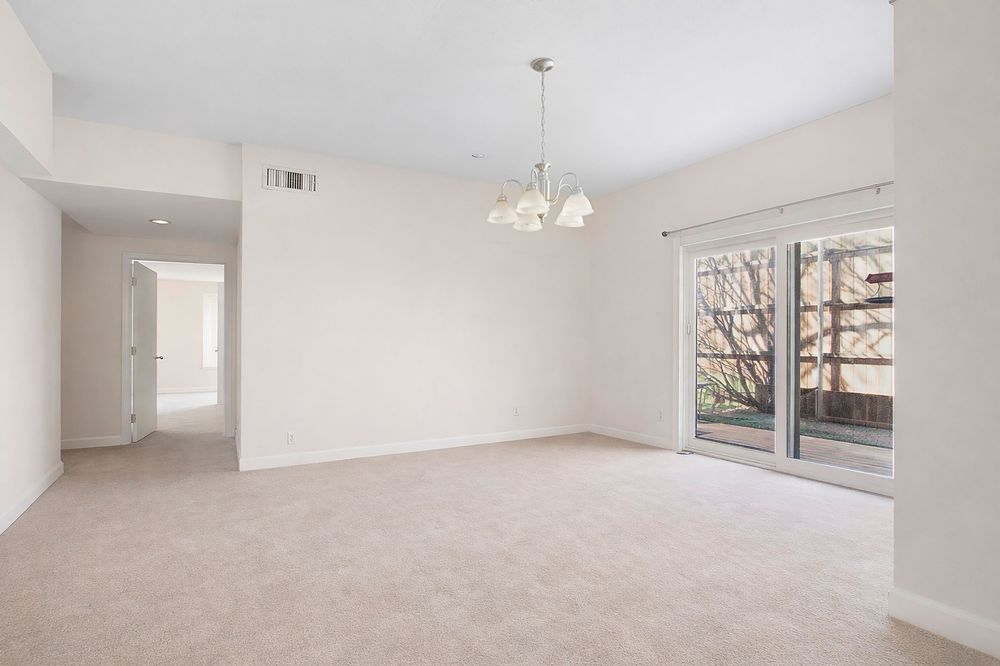 Bright empty living room with beige carpet, white walls, chandelier, sliding glass doors, and a hallway leading to additional rooms.