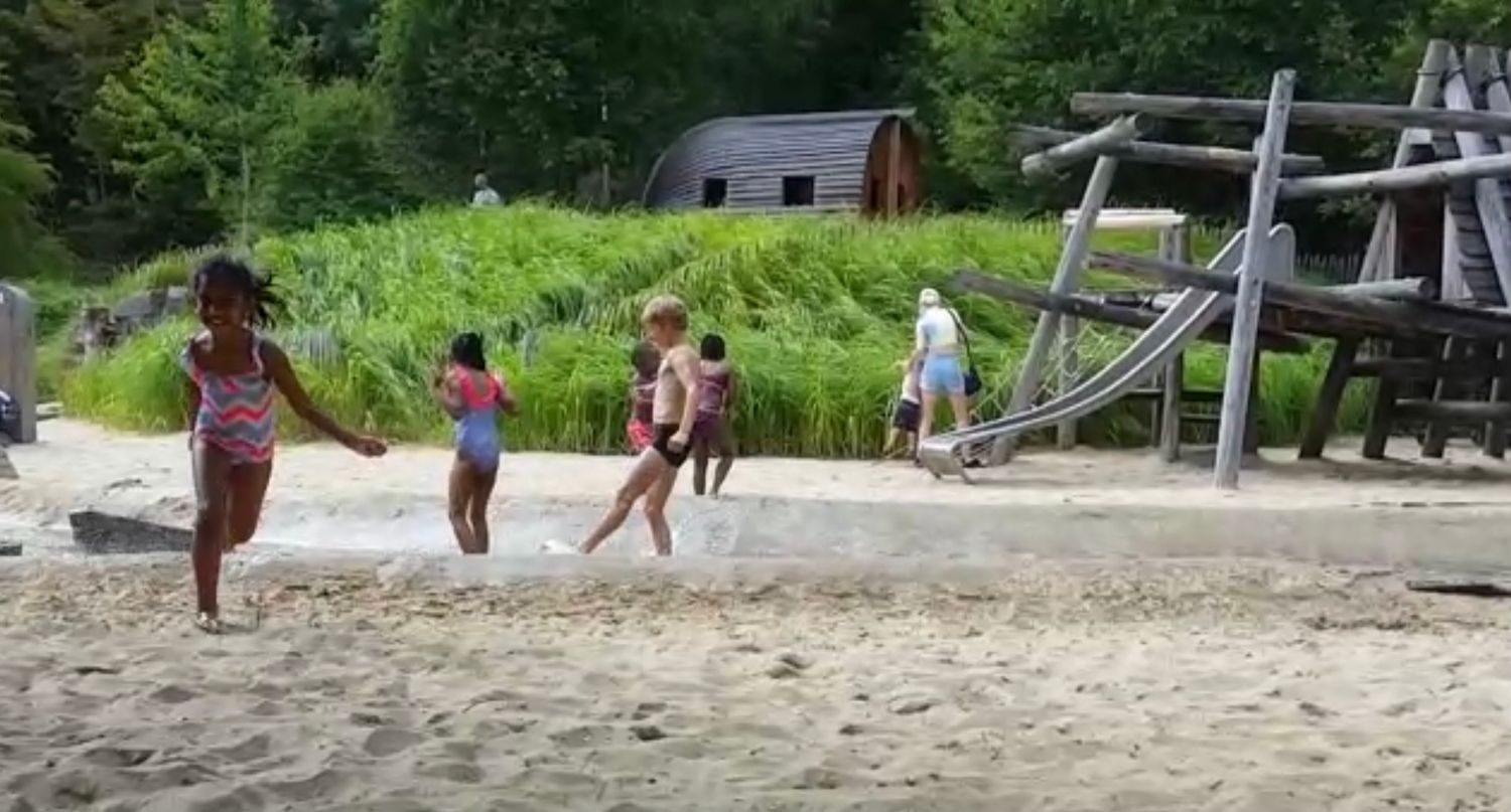 Several kids run and play together on a sandy beach near wooden playground equipment on a summer day.