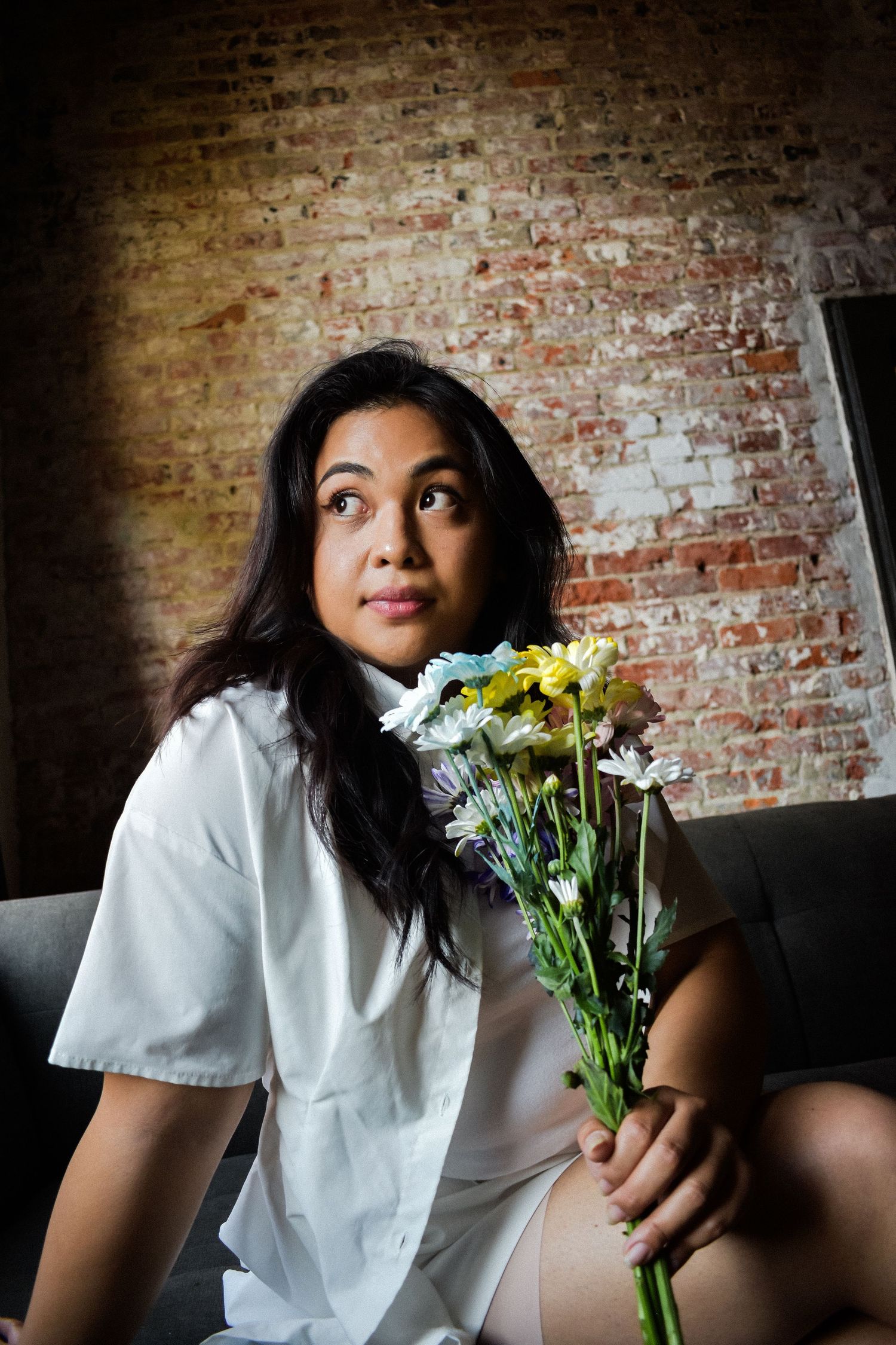 Person in white shirt holds fresh bouquet of yellow and white flowers while sitting against rustic brick wall. Seattle photographer