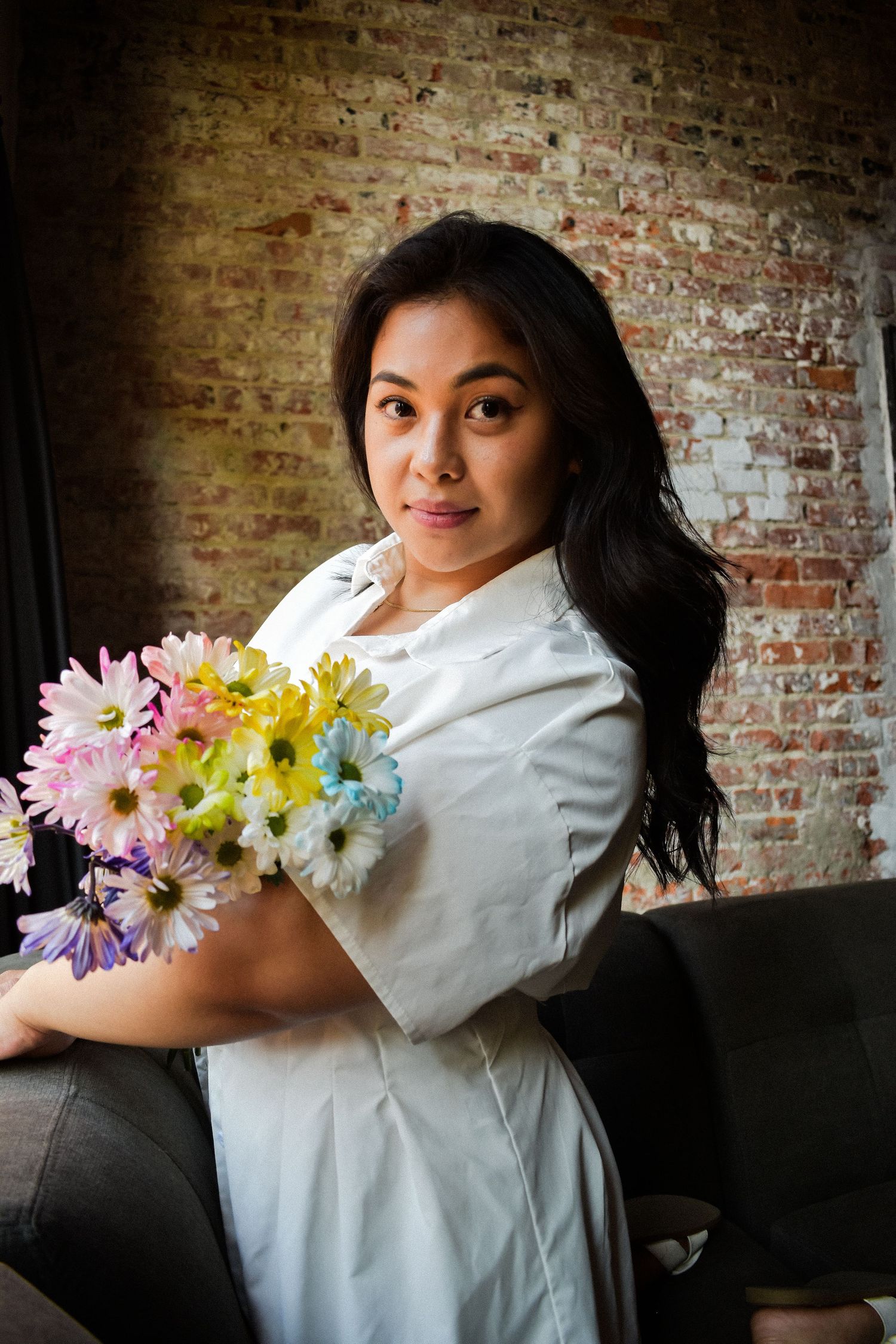 A person in a white dress holds a bright bouquet of colorful daisies against a rustic exposed brick wall. Seattle photographer