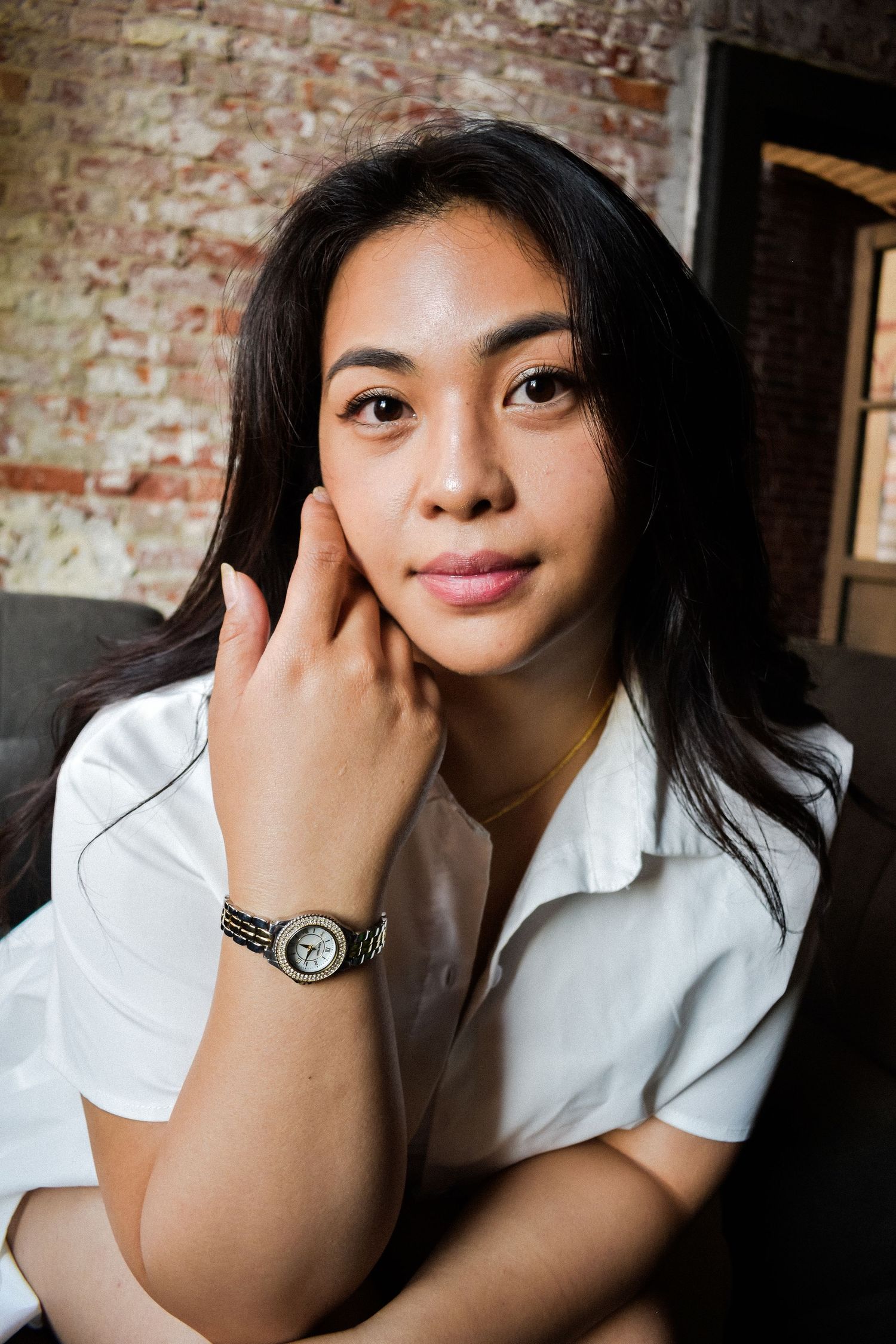 Portrait of a young Asian woman in a white shirt with long dark hair and a wristwatch against a brick wall background. Seattle photography