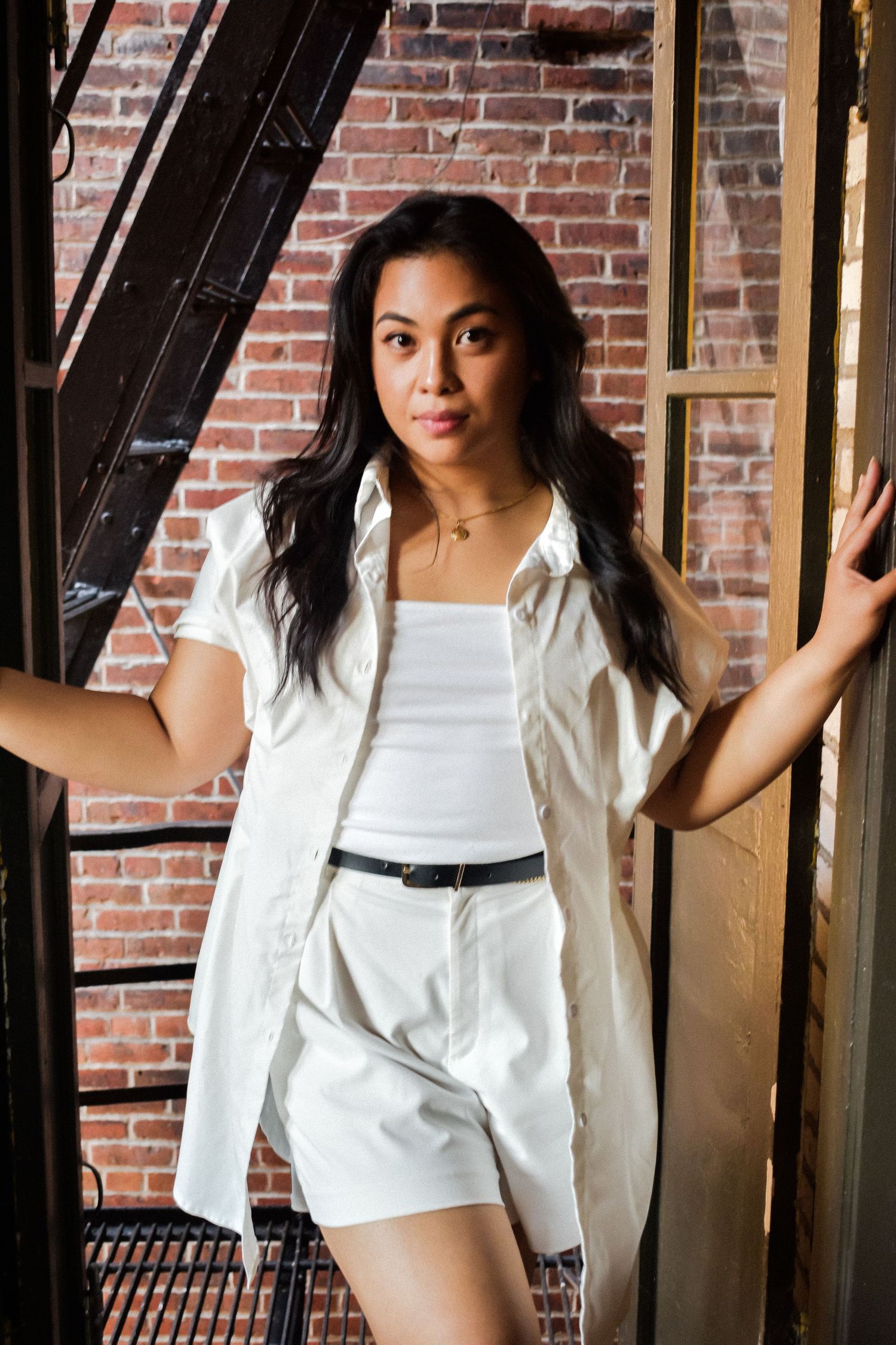 A woman in an all-white summer outfit poses on an industrial fire escape with exposed brick wall background. Seattle branding photography