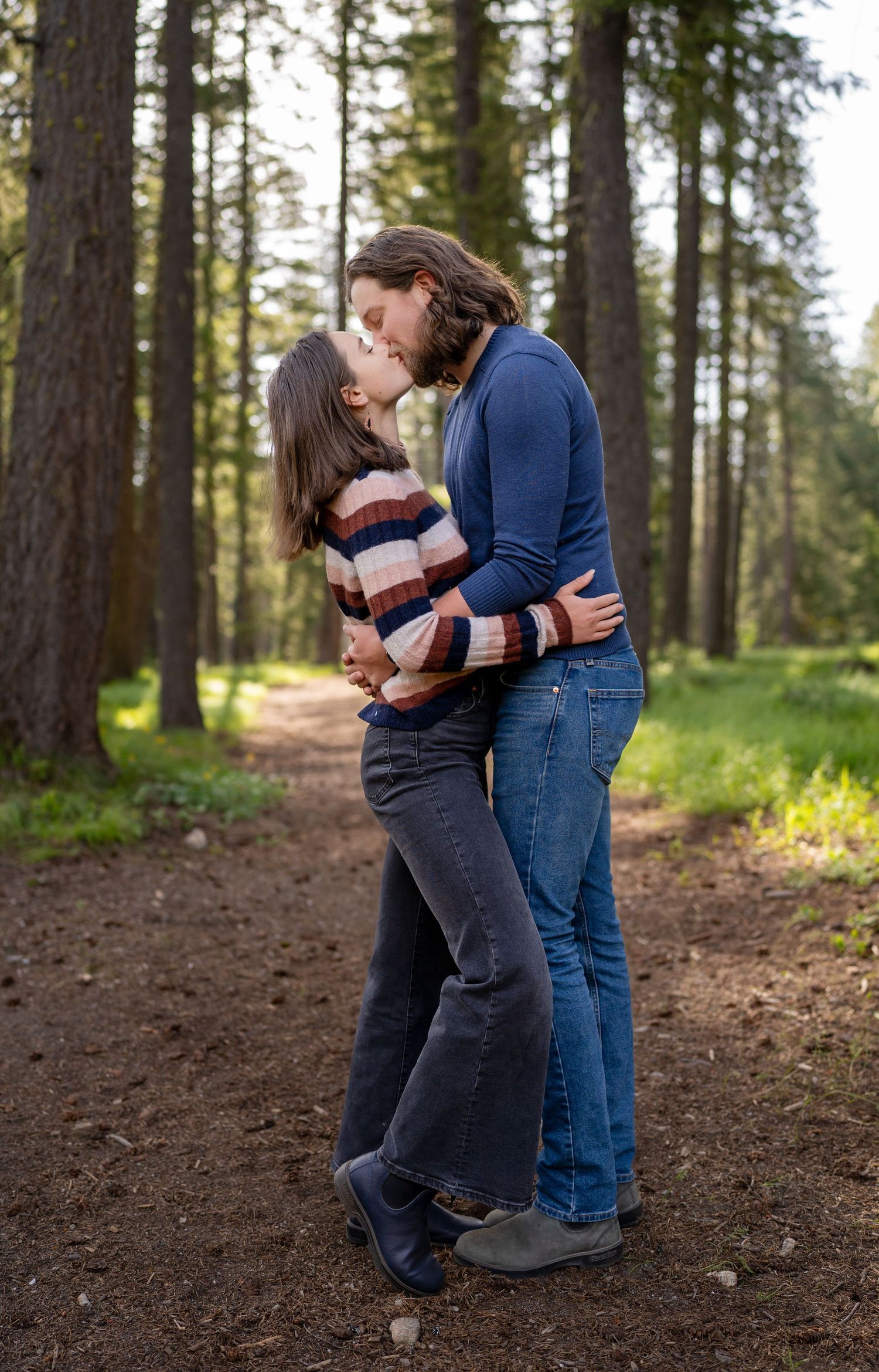 A tender moment between a couple on a forest trail surrounded by tall pine trees.