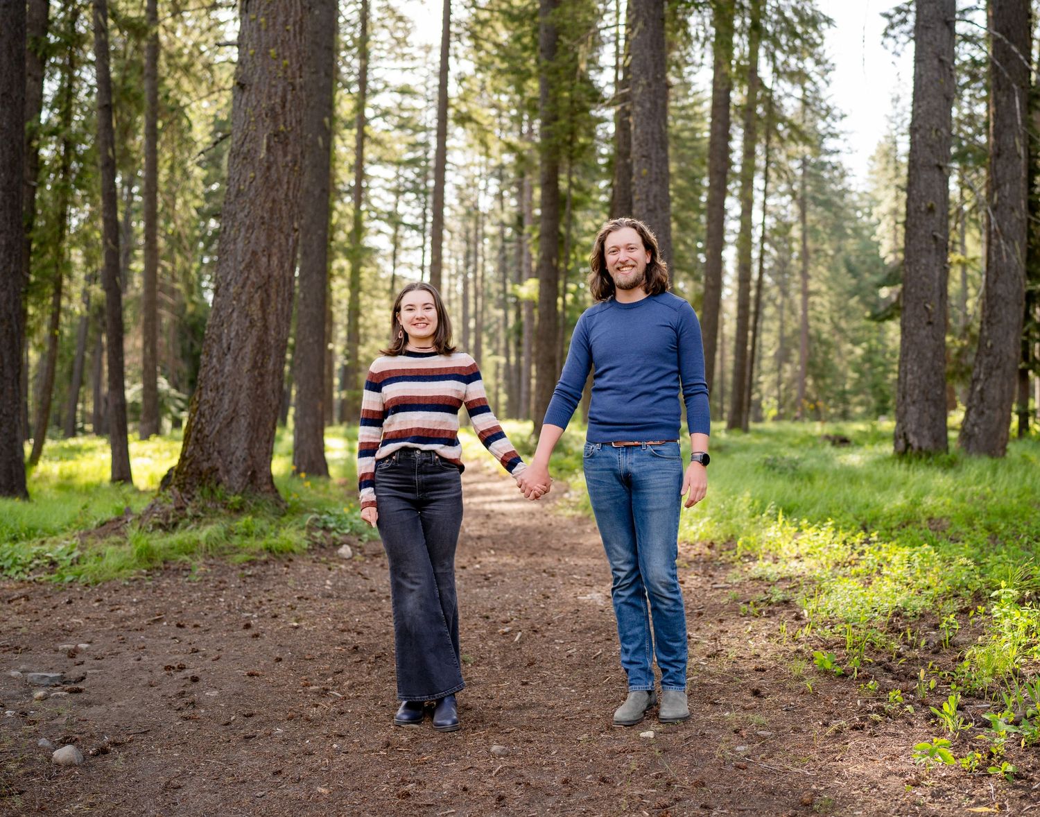 Two people walking together on a forest trail surrounded by tall pine trees.
