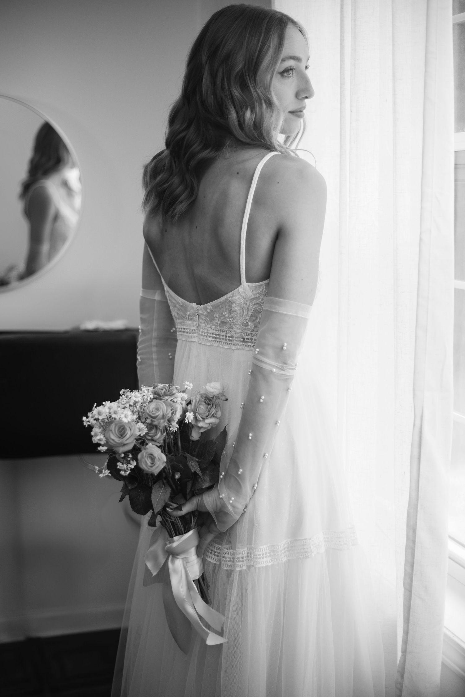 A black and white photograph shows a bride in a delicate lace gown holding a bouquet while gazing out a window.