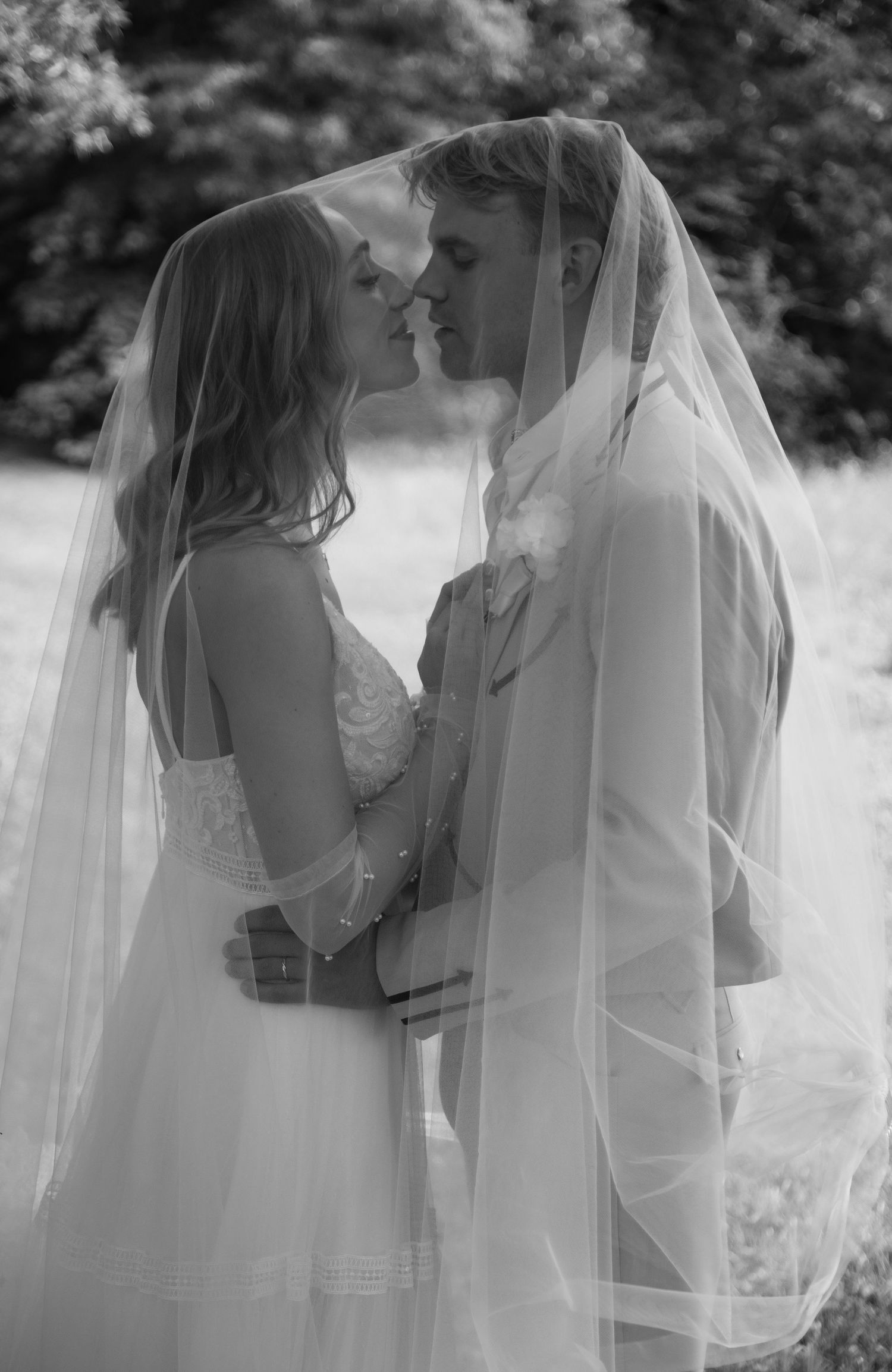 A romantic black and white wedding photo showing a tender moment between a couple under a flowing veil outdoors.