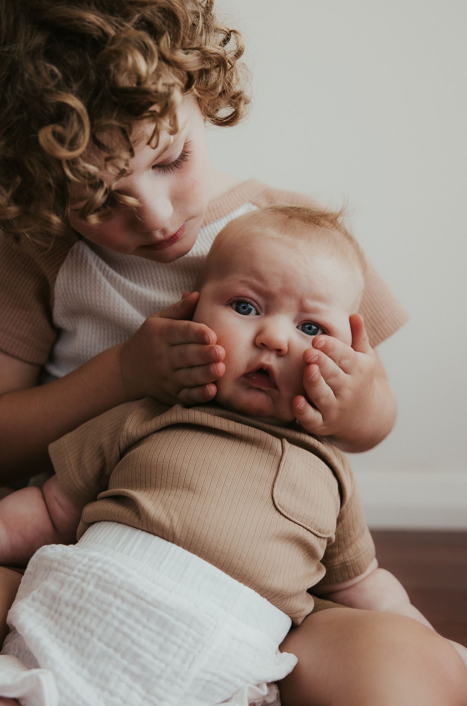 Siblings share sweet moment as older child hugs baby in matching neutral outfits.