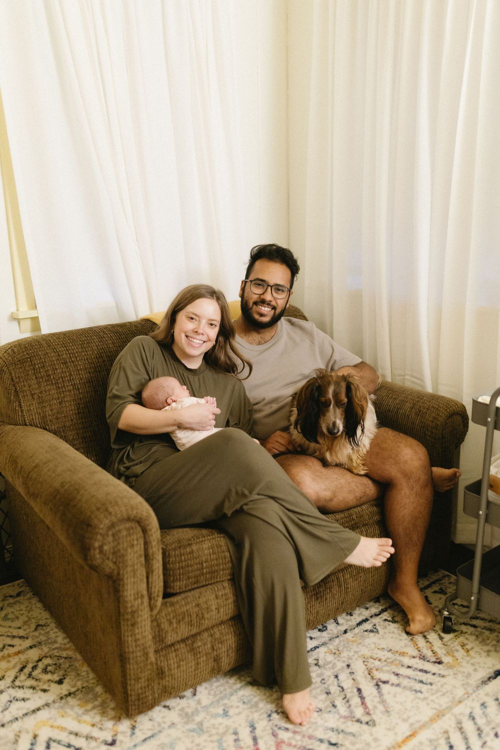 Family relaxing on brown couch with their dog and newborn baby in cozy living room.