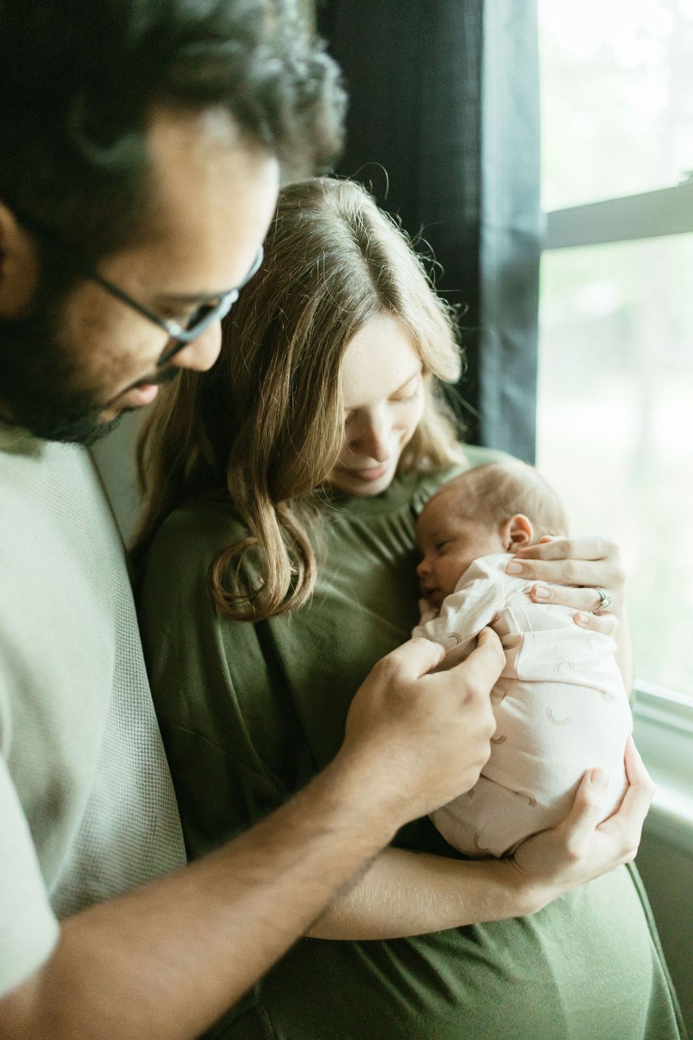 Intimate family moment by window as parents cradle their newborn baby.