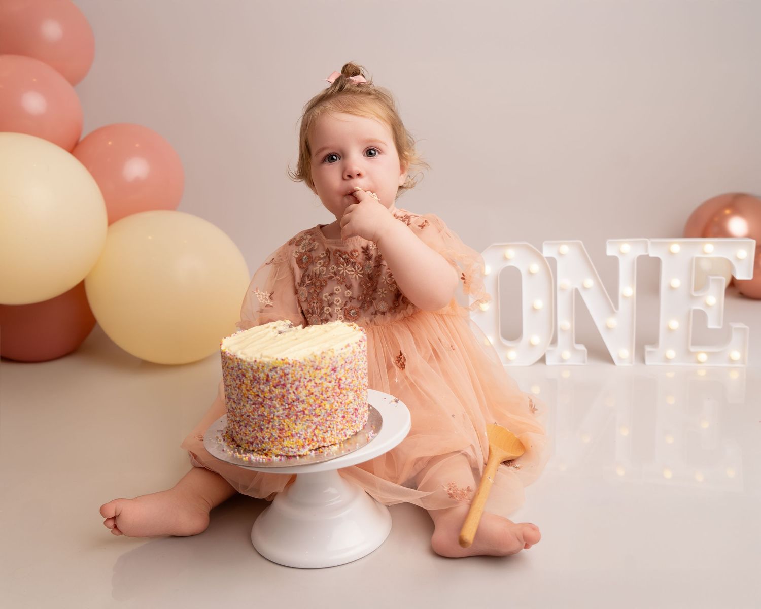A toddler in a peach tulle dress sits beside their first birthday cake with ONE letters in the background.