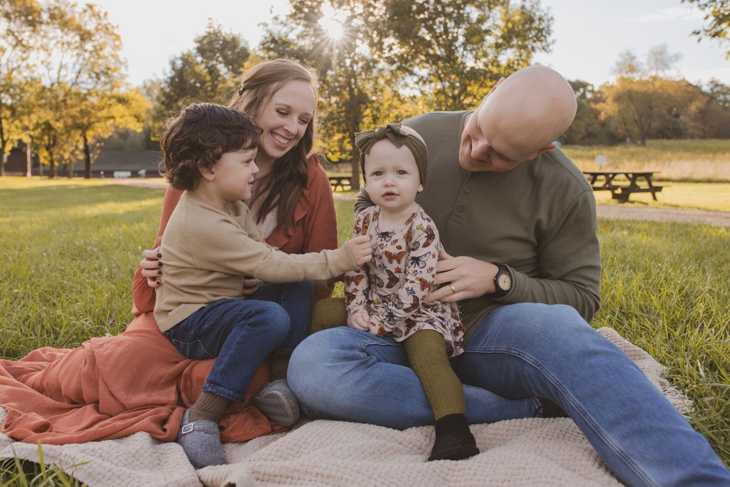 A family sits together on a blanket in a sunny park during autumn while sharing a joyful moment together.