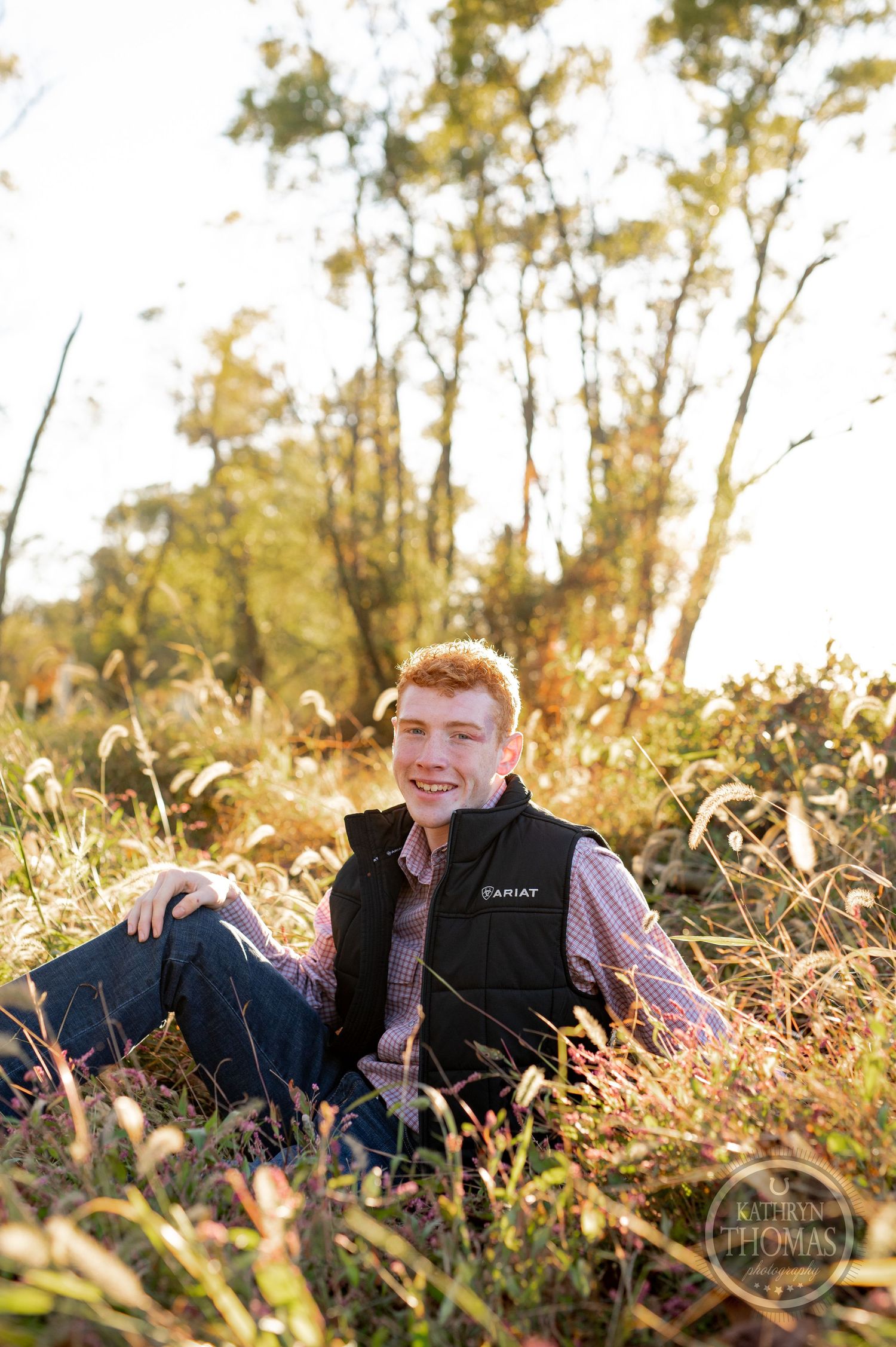 Portrait of a person sitting in autumn foliage wearing a black vest and checkered shirt at sunset.